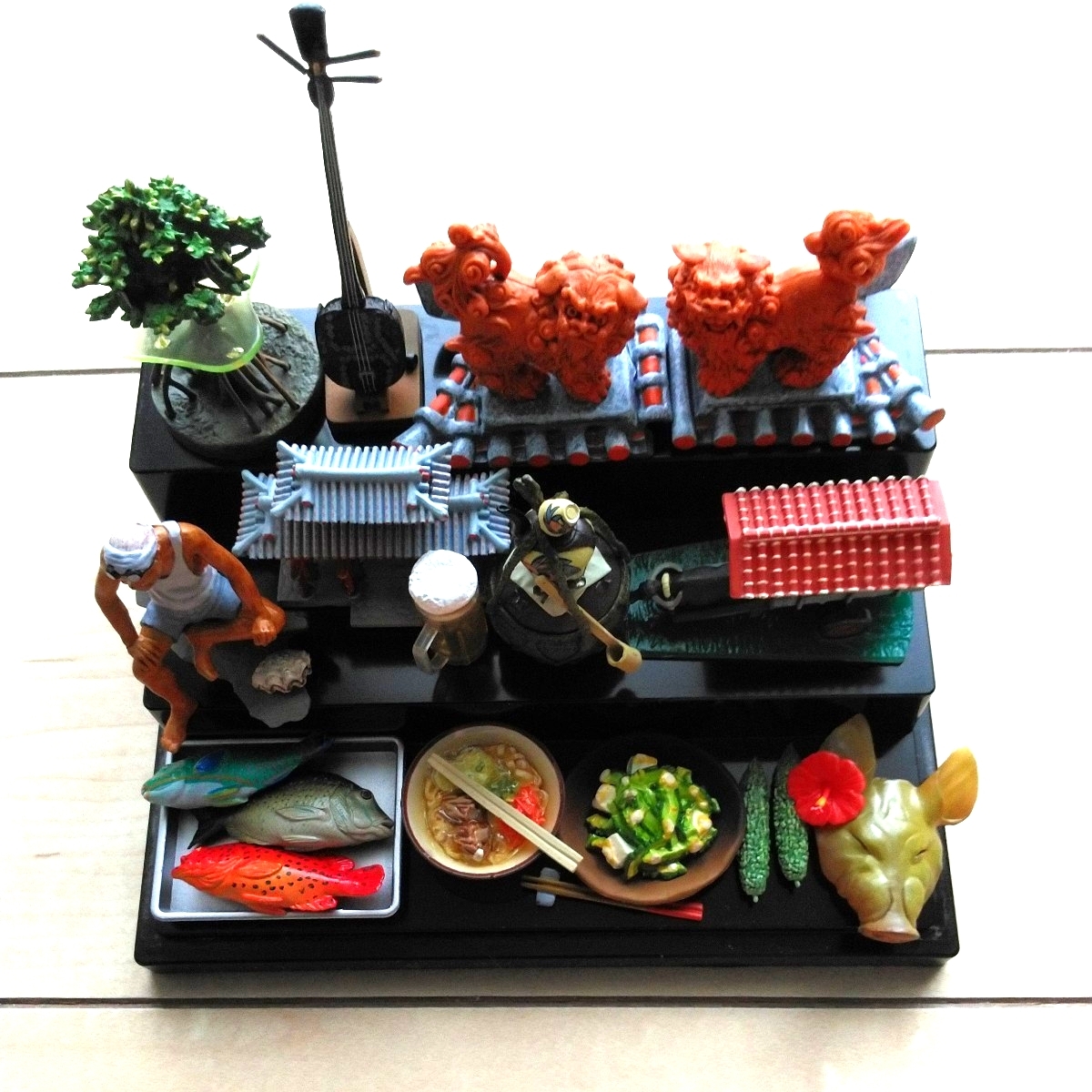 ■『沖縄県の風物詩Figure』13点一括Set。黒色Plastic製雛壇に透明Case付。守礼門・瓦屋根上の魔除けシーサー・沖縄そば・石垣島牛車etc.拍卖